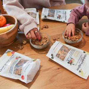 Two people eating granola from bowls on a wooden table with packaging around.