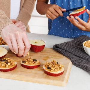 People preparing apple slices with peanut butter and granola on a wooden cutting board.