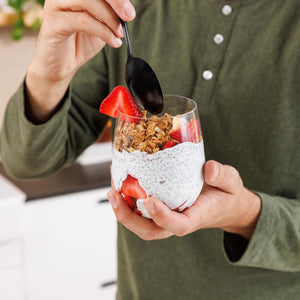 Person holding a glass of chia pudding with strawberries and granola, using a spoon.