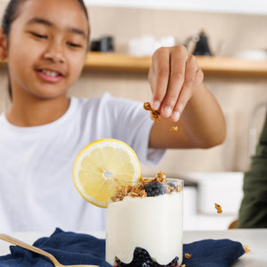 Person adding toppings to a glass of yogurt with a lemon slice and blueberries.