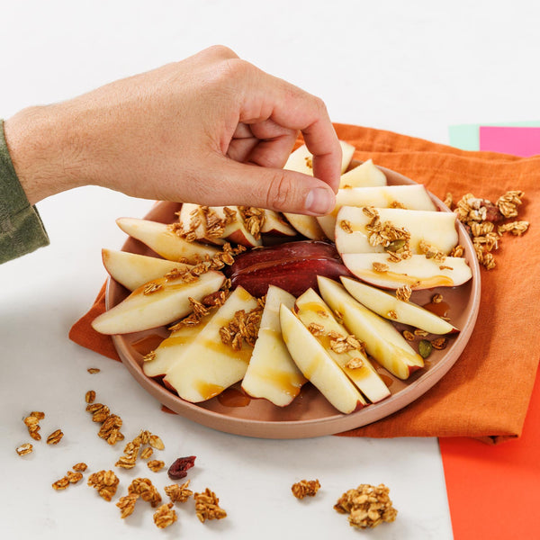 Hand reaching for apple slices with granola on a plate, placed on an orange napkin.