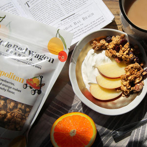Breakfast scene with granola bowl, orange, and Little Red Wagon granola package on a table.