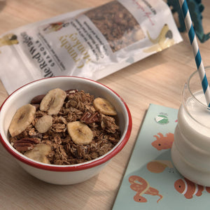 Bowl of granola with bananas and a glass of milk on a table with a newspaper and children's book.