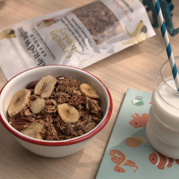 Bowl of granola with bananas and a glass of milk on a table with a newspaper and children's book.