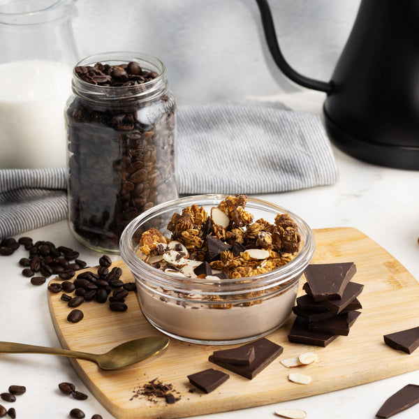 Glass bowl of granola with chocolate pieces on a wooden board, surrounded by coffee beans and a jar of chocolate chips.