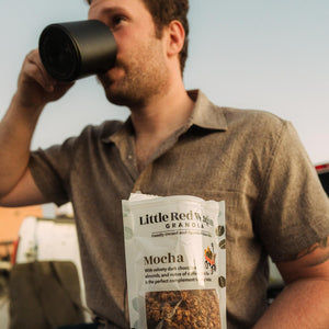 Man drinking coffee from a black mug with a 'Little Red Wagon' granola bag in the foreground.