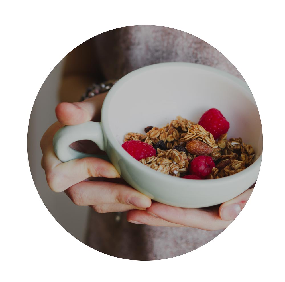 Person holding a white bowl filled with granola and raspberries.